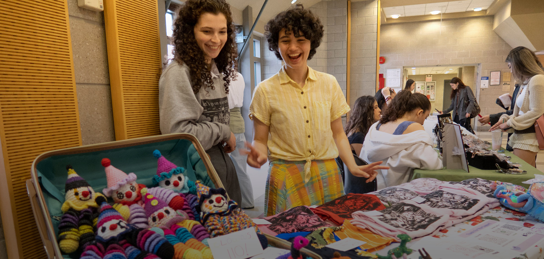 Two female students standing behind a table of crafts they are selling such as yarn puppets and screenprinted tshirts.