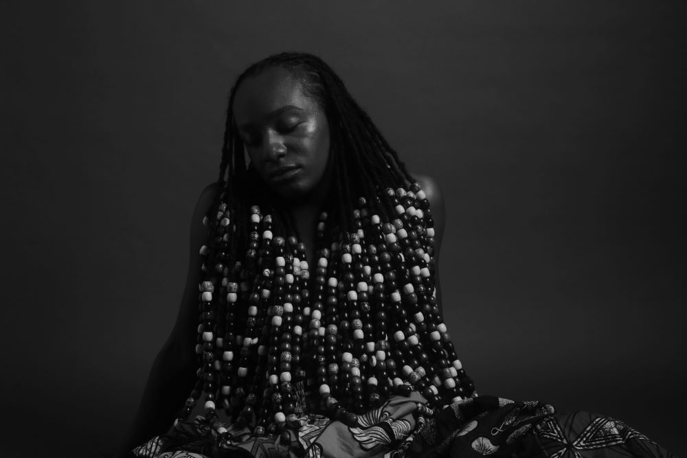 At the center of a black and white horizontally oriented photograph is a figure seated on the floor or ground. The eyes are closed, head tilted to the side, long hair is styled in dreadlocks that are adorned in large beads shaded from light to dark. The braids are draped to completely cover the upper body and torso with only naked arms that are placed behind for support slightly exposed. A little bit of Ankara patterned textile covering the lower body visible at the bottom, completing the image.