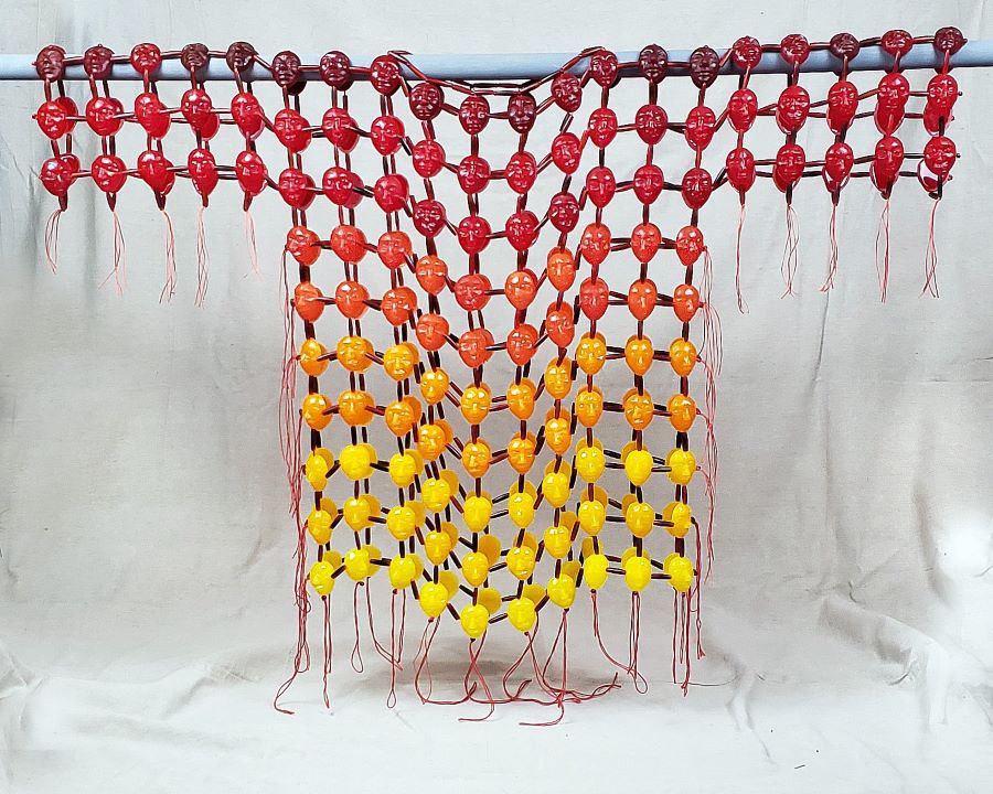 A war shirt made of large yellow, orange, red and maroon colored beads shaped like faces, sparsely strung in a lattice pattern. The garment is reminiscent of armour. The maroon starts at the top, and the color shades gradually change to yellow at the bottom of the shirt. 