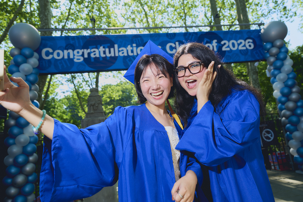 two students in blue regalia posing with a 2026 commencement banner behind them