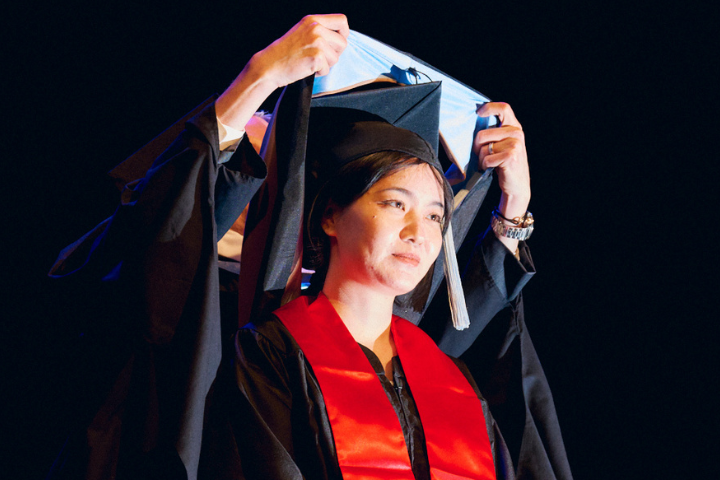 student wearing red sash signifying China receives master's hood from her department chair at commencement ceremony