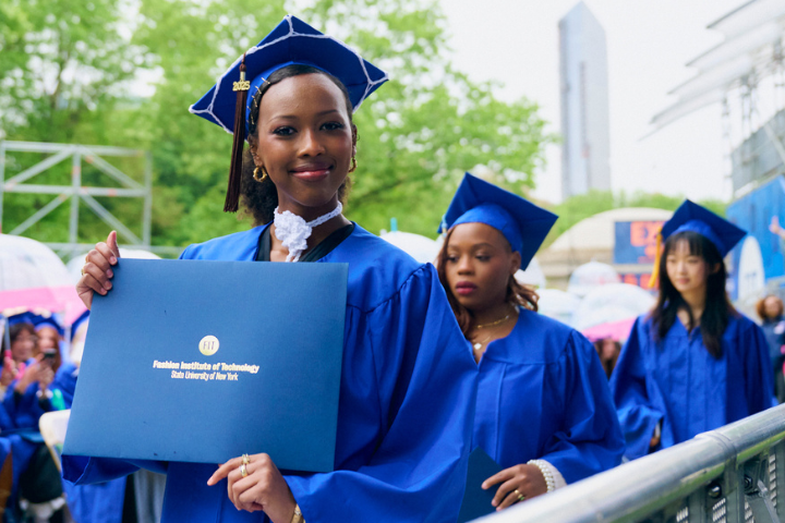 students in blue regalia walk in front of classmates in regalia; one of the students poses and holds diploma folder