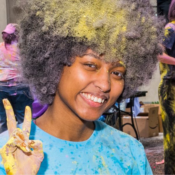 a student smiling, and holding up her hand in a "V" gesture. Her hand, hair and skye-blue t-shirt are spotted in yellow paint-powder the picture seems to be taken during a Diwali celebration event  