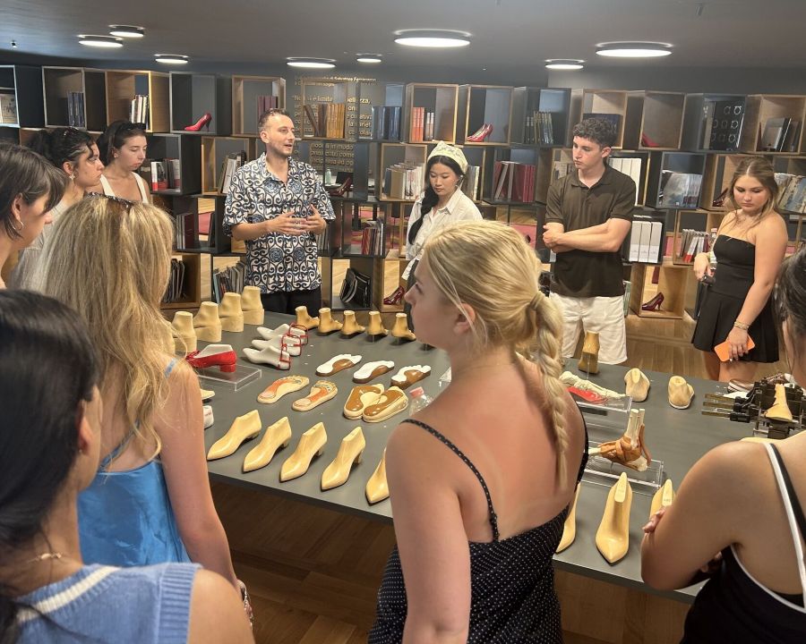 Instructor is speaking to a group of students gathered around the table with the shoemaker's forms, prototypes of the foot beds, and other footwear production tools displayed on the table