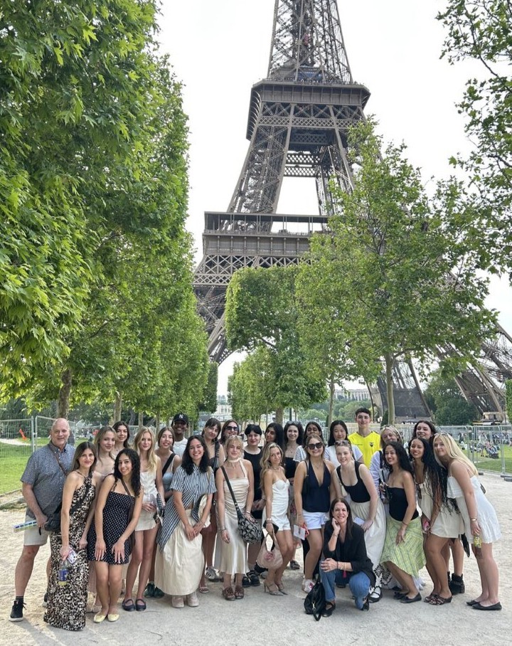 Group of students posing for a photo on a park path lined with green trees and the Eiffel Tower in the background