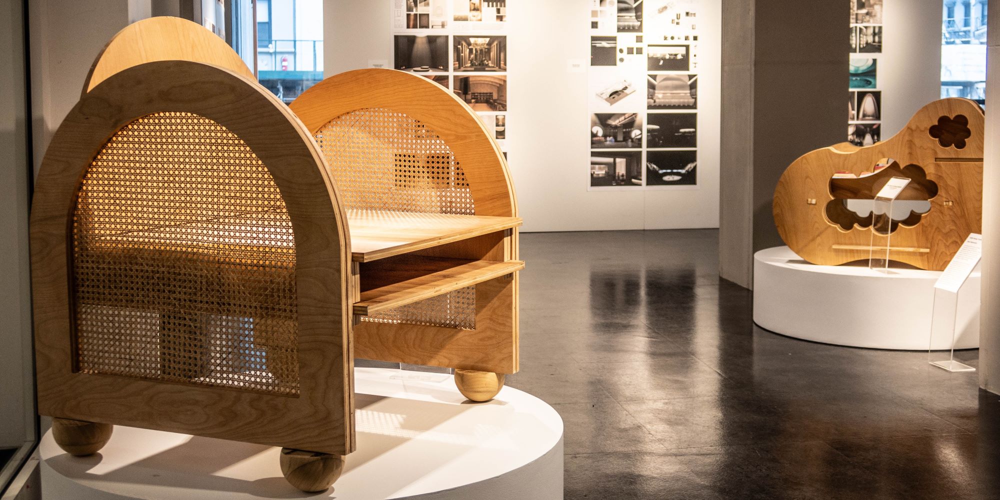 Two wooden arm chairs displayed on the round platforms. both pieces of furniture feature natural wood and rounded elements. Renderings of interior design work on the walls