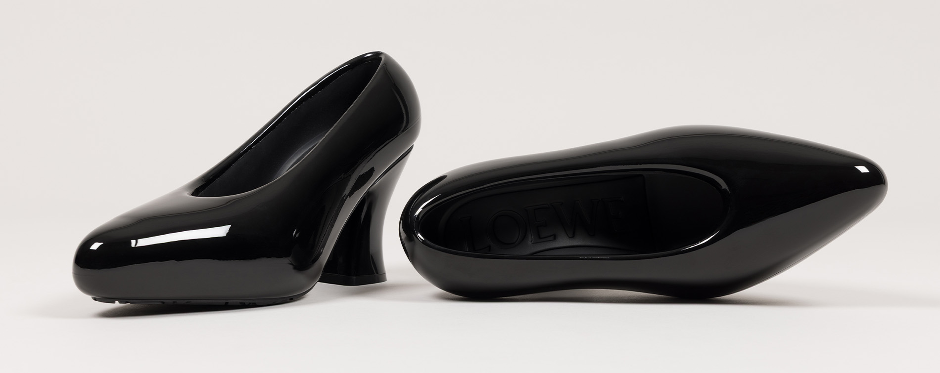 Two glossy black sculptural high-heeled shoes displayed on a white surface, one upright and one on its side showing the branded sole.
