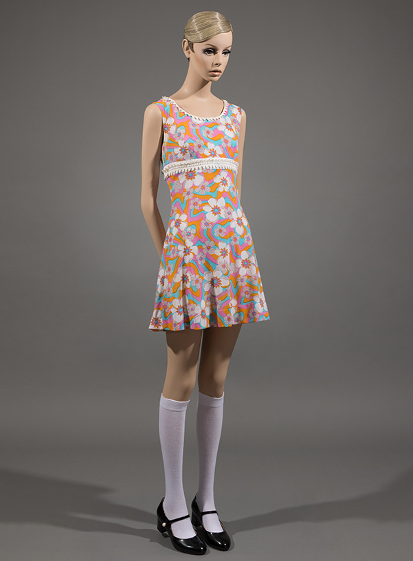 twiggy mannequin wearing sleeveless short length dress with a pink flower print, styled with white knee high socks and black mary jane shoes
