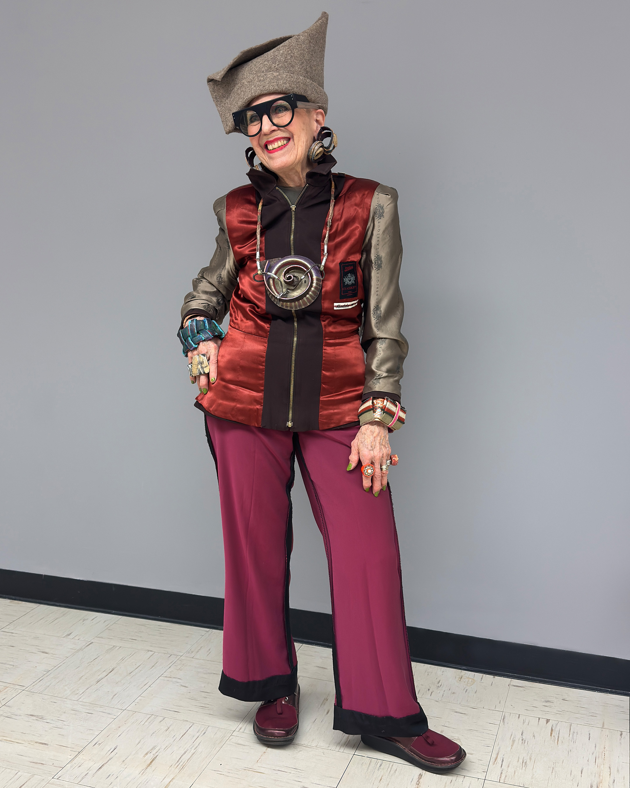 Debra wearing a red satin jacket styled with magenta pants with black side seams, black shoes, and a gray hat