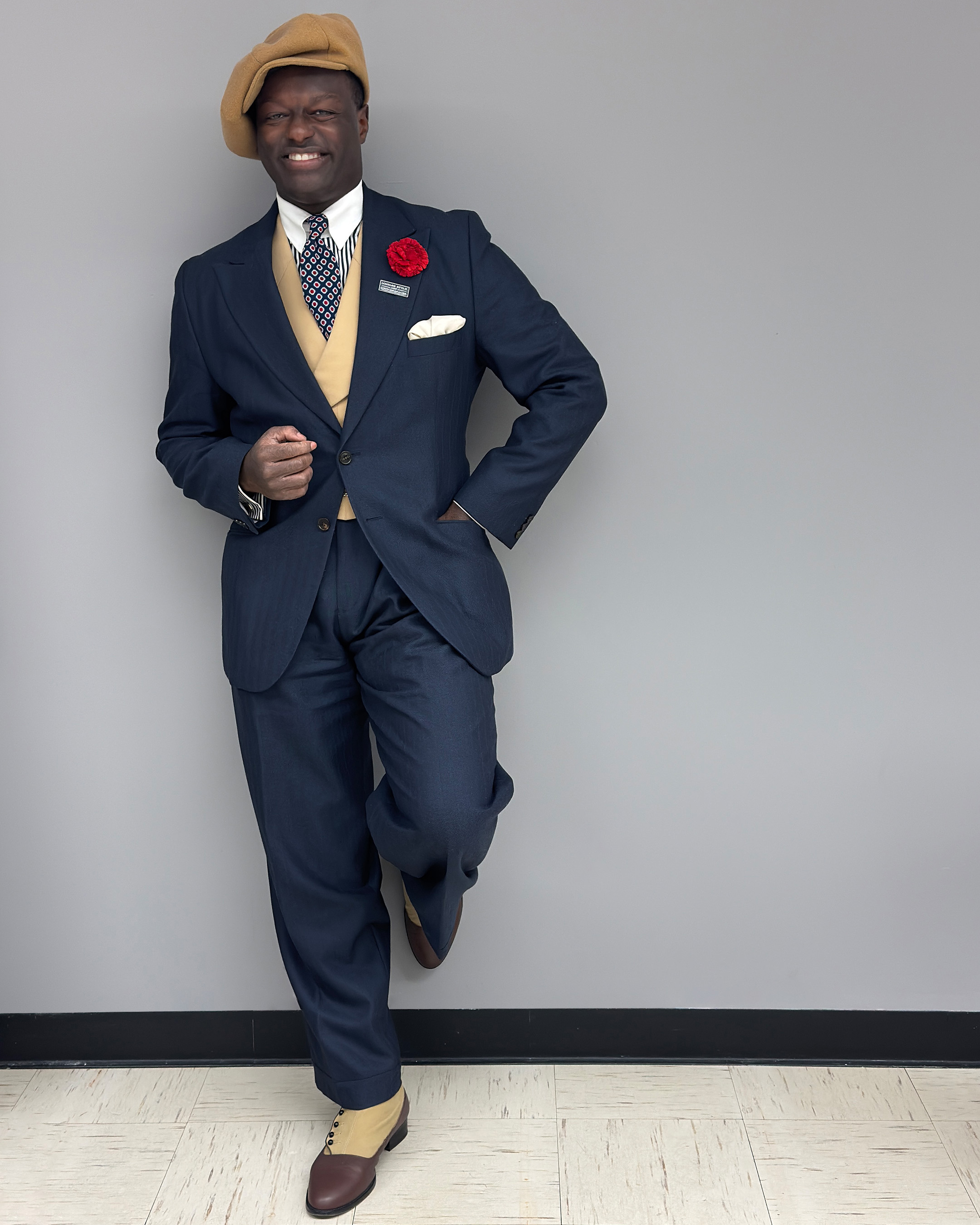 Dandy wearing a tan newsboy hat with a navy, peak label suit styled with a tan waist coat, blue, red, and white medallion tie, and two toned brown button boots