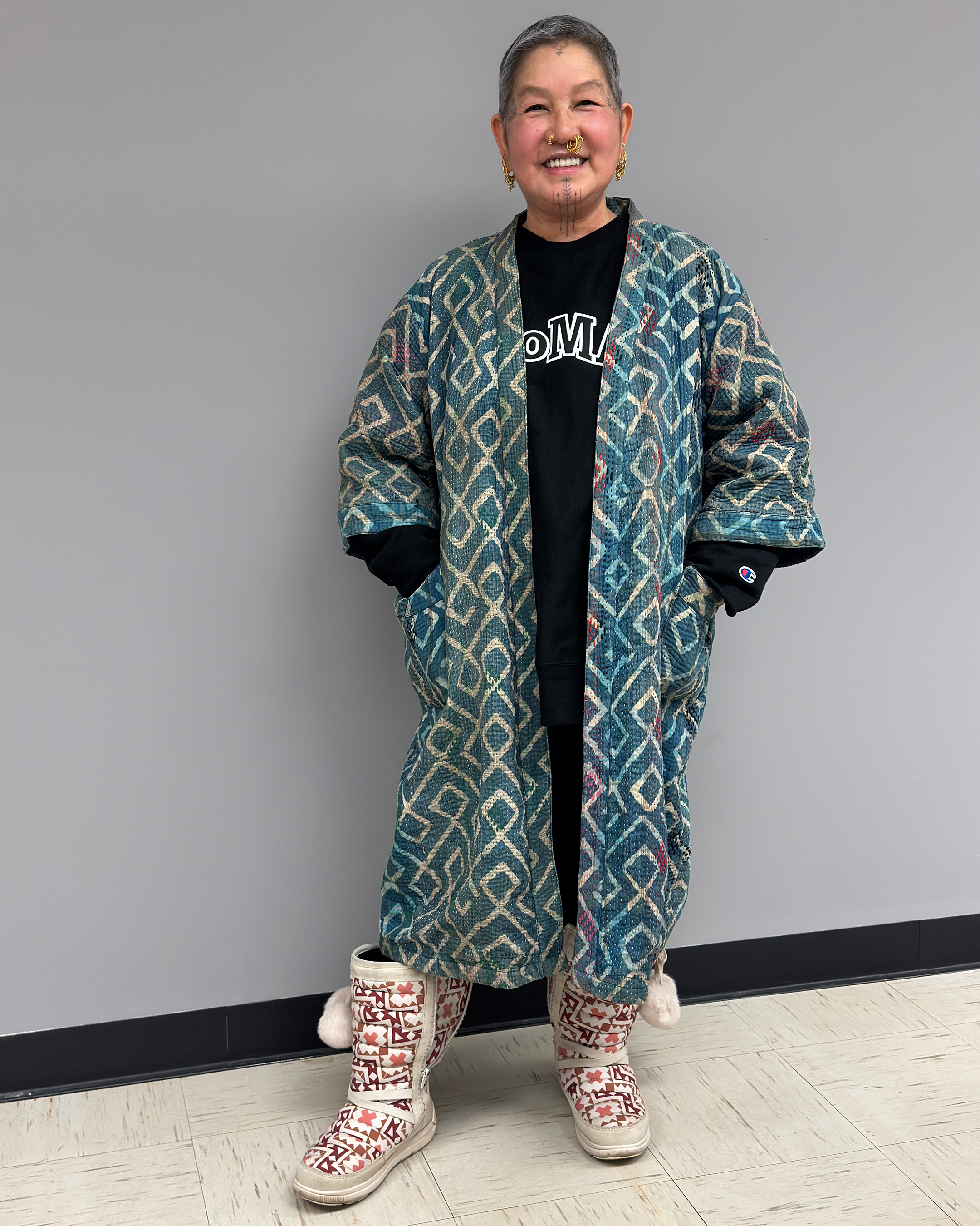 Cynthia with multiple nose piercings and face tattoos, wearing a long, open indigo kantha quilt featuring a geometric pattern in teal, cream and rust over a black MoMA sweatshirt. She is also wearing cream and pink Manitoba Mukluk boots with white pom poms