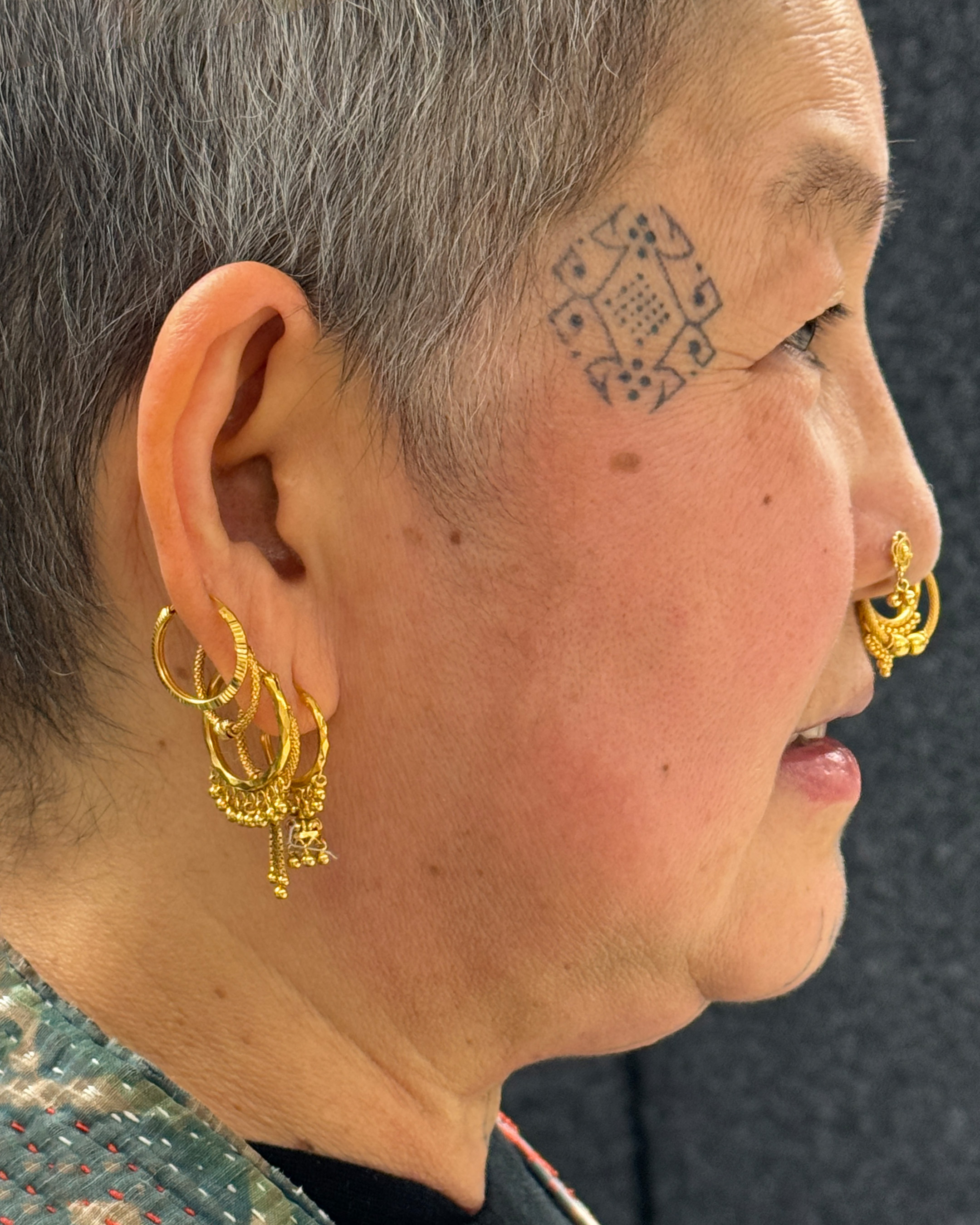 Cynthia's side profile, showcasing her face tattoo, a traditional geometric diamond motif at her temple, and an stack of intricate gold filigree hoops at her ear, finished with a matching gold nose ring.
