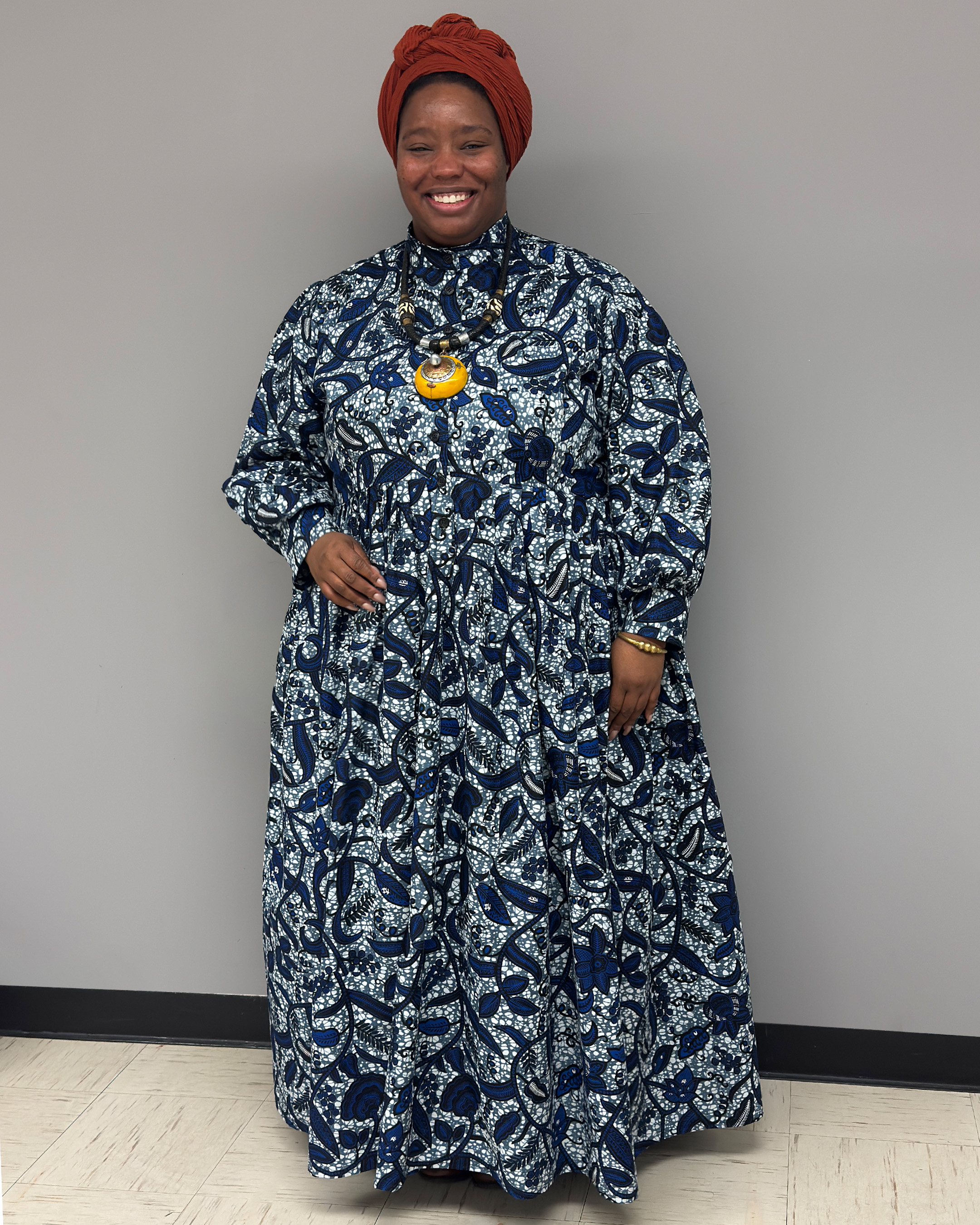 Cheyney, a Black woman, wearing a long sleeve, long blue print dress with a chunky yellow necklace and a rust colored head wrap
