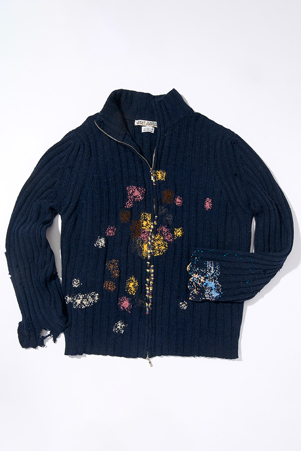 Close up of a black top with embroidered safety pins and x-marks on the shoulder. The top is laid flat, with a spool of blue yarn with a needle is placed on top