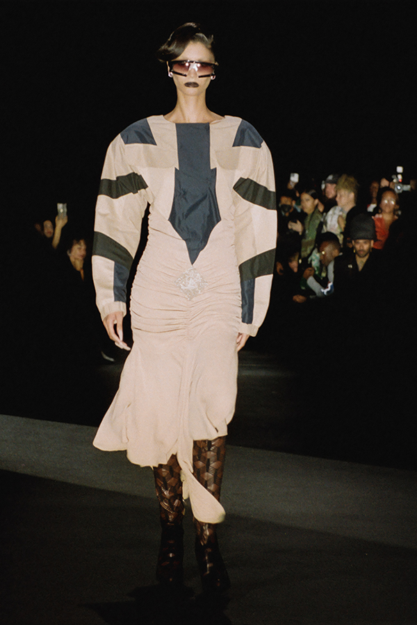 Model on runway in taupe and blue colorblock dress with broad shoulders and fitted skirt