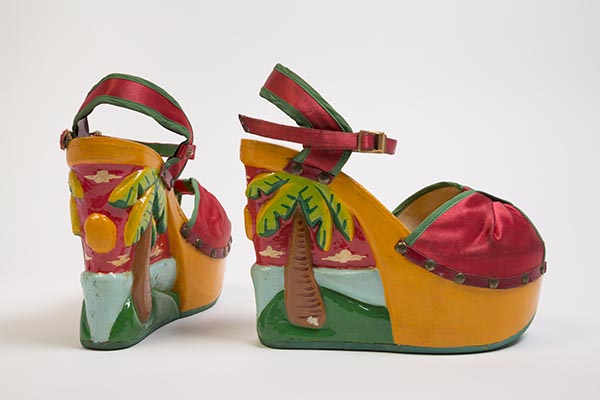 Red ankle strap platform wedge open toe sandals in synthetic satin with hand painted and carved relief design on polyurethane platform base of palm trees and tropical beach scene in red, yellow, blue, green, orange, brown and white, upper front panel cinched at CF and held in place with self fabric band, green fabric bias binding at finished edges, upper shoe attached to base with tarnished gold metal nail heads and leather trim with fluted copper metal stud at inside strap, copper metal buckle at ankle strap closure