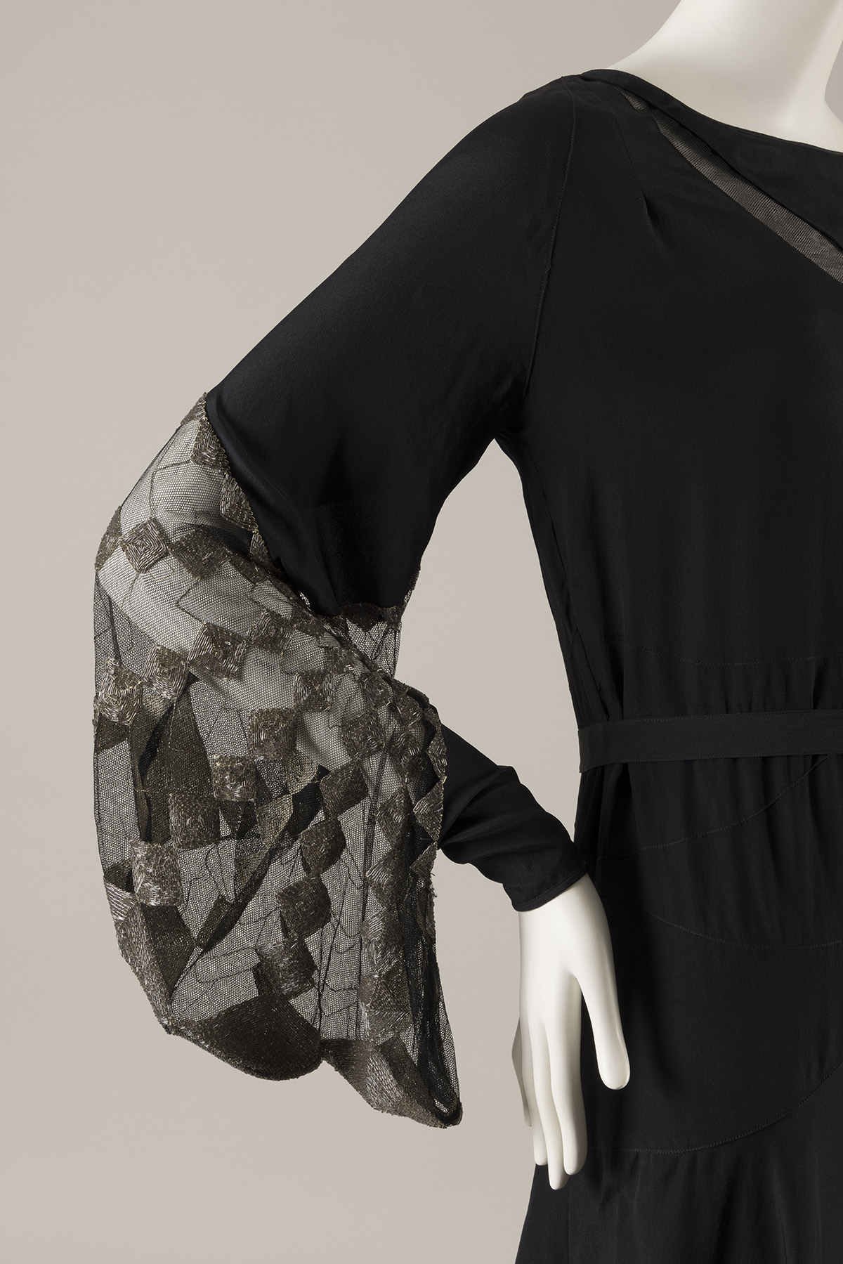 A black silk dress with a straight silhouette. The lower portion of the dress’s long sleeve features an insert of black net embroidered with silver beads in a geometric pattern.