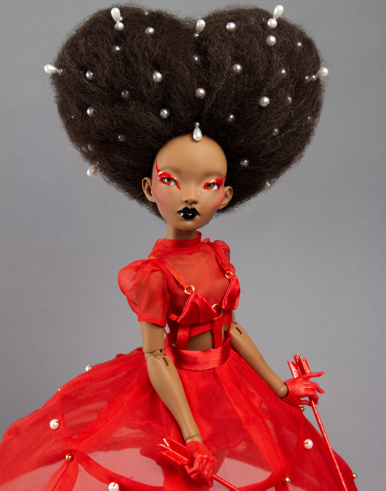 Black doll wearing a red dress and an afro shaped into a heart with embedded pearls. 